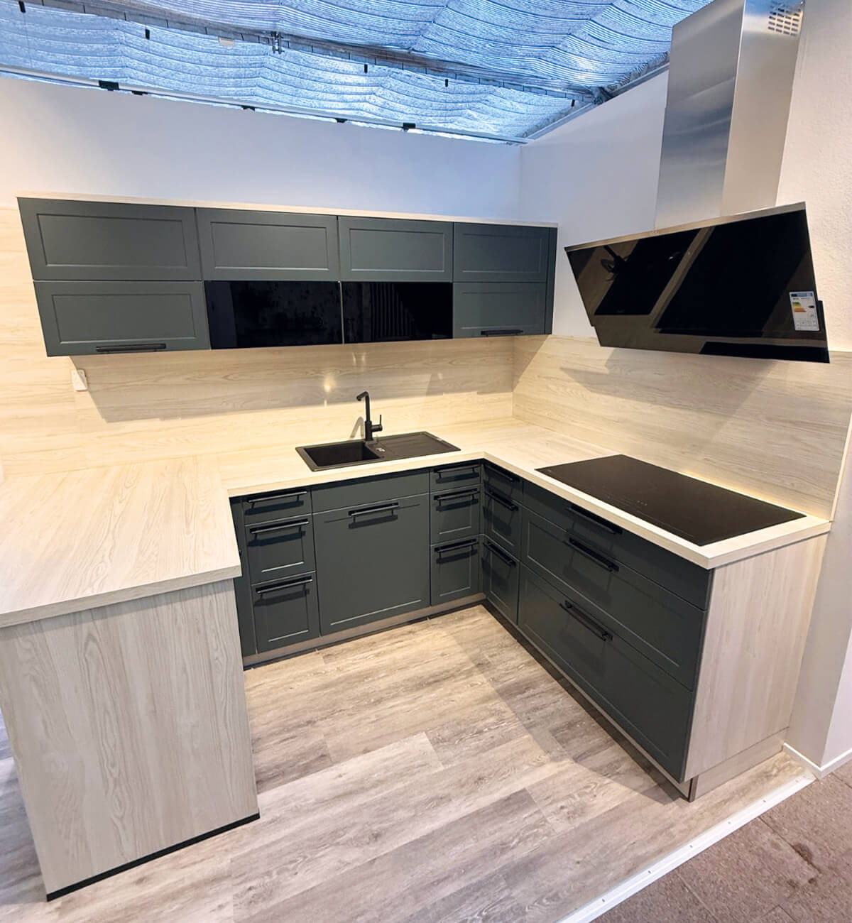 Moderne L-förmige Küche mit dunklen Fronten, hellem Arbeitsplatten- und Wandbereich, integriertem schwarzen Spülbecken und Kochfeld sowie schräger Dunstabzugshaube